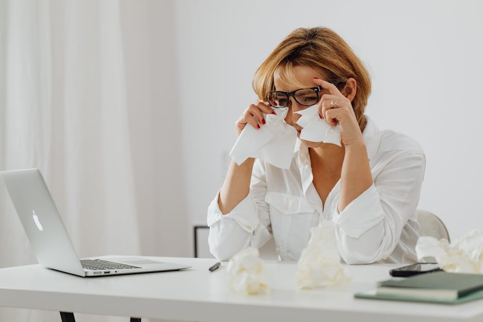 A woman in glasses is wiping tears while seated at a desk with a laptop and tissues.