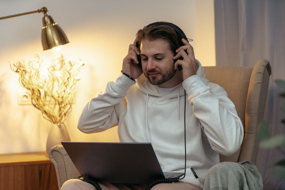 A bearded man in a hoodie enjoys music with headphones while working on a laptop.