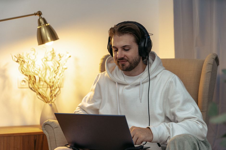 Casually dressed man on a chair using laptop with headphones in cozy interior setting.