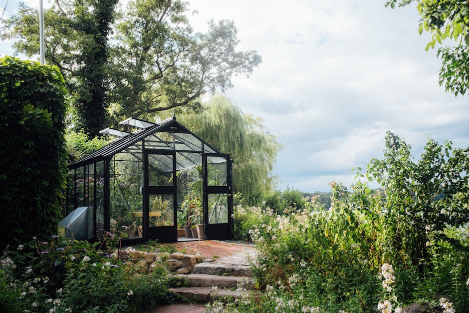 A picturesque greenhouse nestled in a vibrant garden surrounded by greenery and trees under a bright sky.