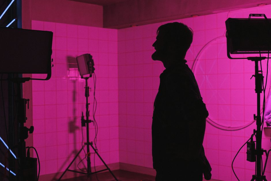 A silhouetted photographer stands in a pink-lit studio with lighting equipment.