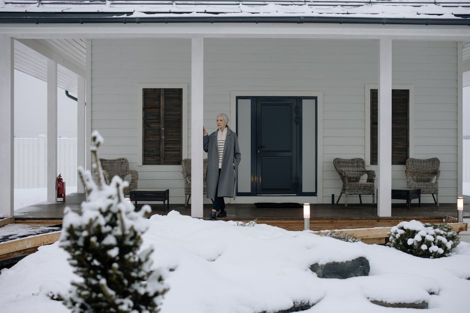 Elderly woman in coat standing on a snowy porch during winter, surrounded by snow-covered landscape.