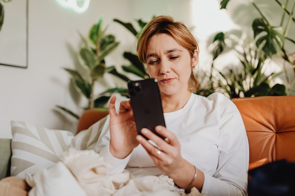 Adult woman using smartphone on a cozy sofa at home, surrounded by plants.