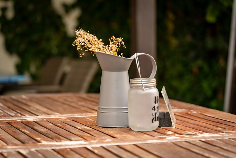 A rustic outdoor table setting featuring a metal pitcher with dried flowers and a jar.